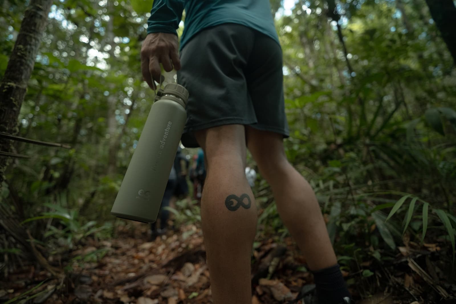 An infinity tattoo on a trail that has no easy shortcuts, fitting. Close-up low-angle view of a hiker's legs on a Pico da Neblina Expedition forest trail, one leg bearing an infinity tattoo, hand holding an olive-green insulated water bottle, with other trekkers visible among dense green rainforest in the background