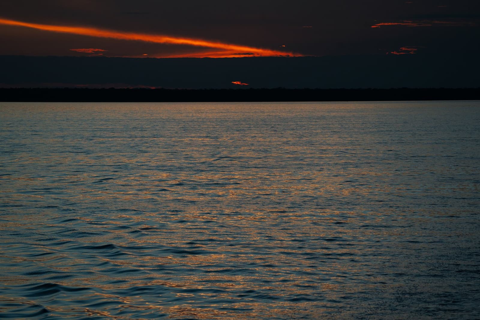 The last light of the day, and the river holds every bit of it. Deep red and orange streaks of cloud glow above a wide dark river at dusk on the Tucano Amazon river cruise, with a low forested horizon silhouetted beneath a near-black sky