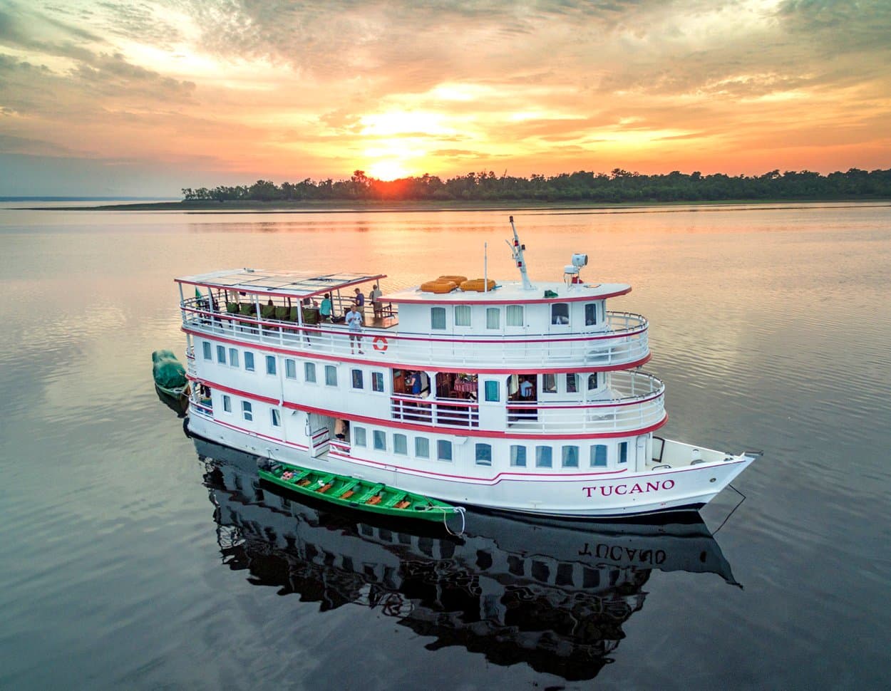 Home for five nights, and the sunsets come with the room. The M/Y Tucano, a white three-deck river cruise vessel, anchored on a glassy dark-water river at sunset, with a green wooden canoe alongside and passengers on the upper deck, forested shoreline silhouetted against an orange sky
