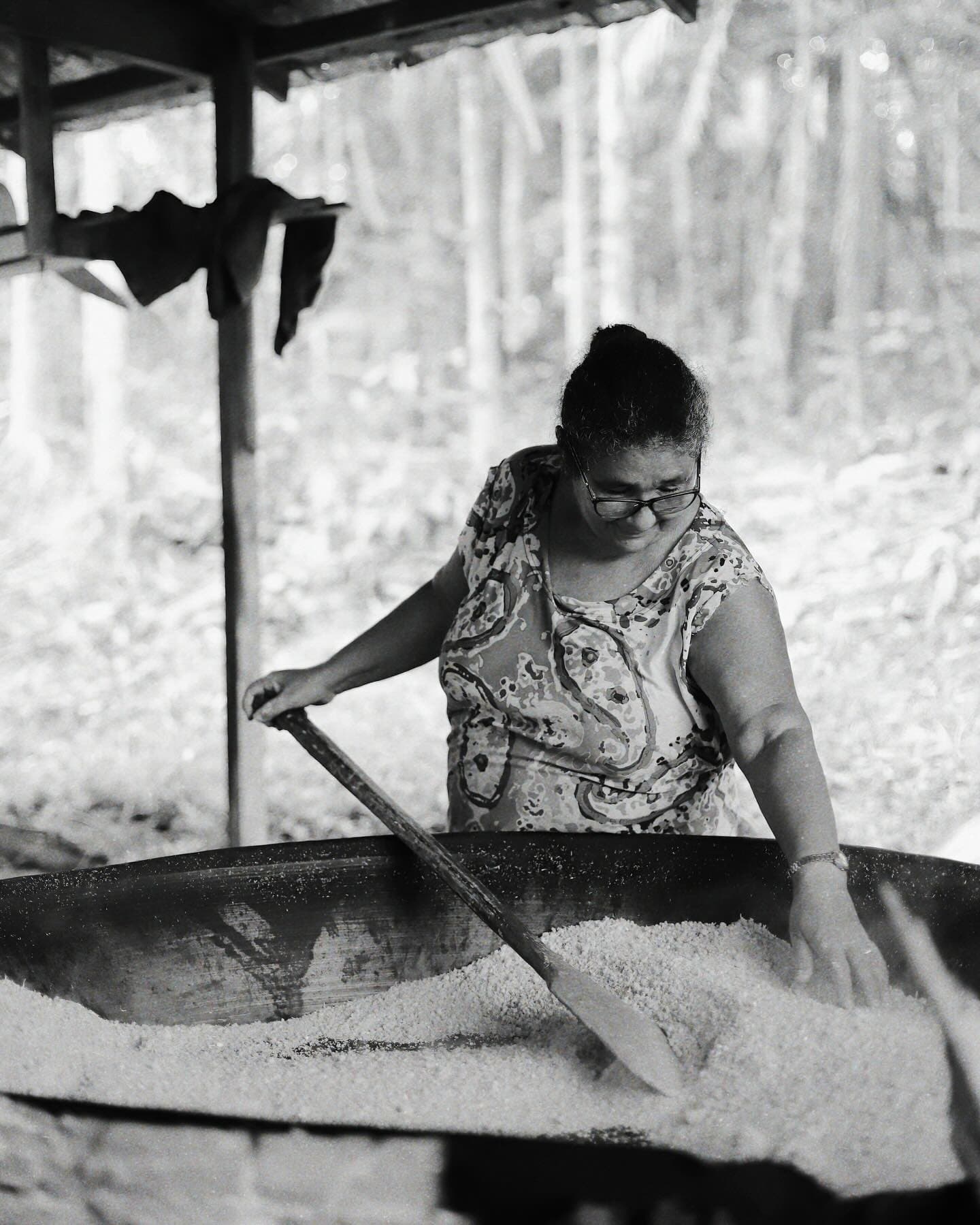 Farinha doesn't make itself, and she's been at this longer than you've been awake. A woman wearing glasses stirs freshly grated cassava in a large iron pan at an open-air flour house during an Amazon Cruise Trip in Brazil, with forest visible in the background