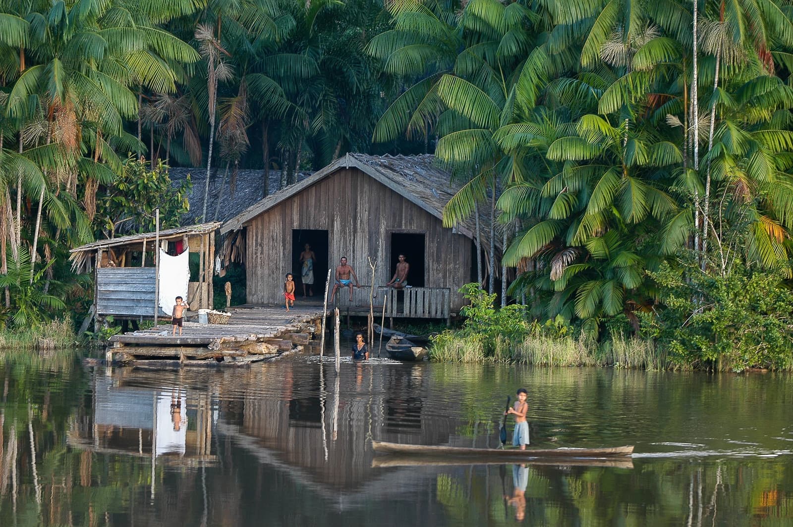 River life here runs on its own quiet clock. A wooden stilt house on an Amazon riverbank surrounded by dense palm forest, with a family on the porch and dock, a person wading at the water's edge, and a child standing in a dugout canoe on the calm river — a glimpse of caboclo life on an Amazon cruise trip in Brazil