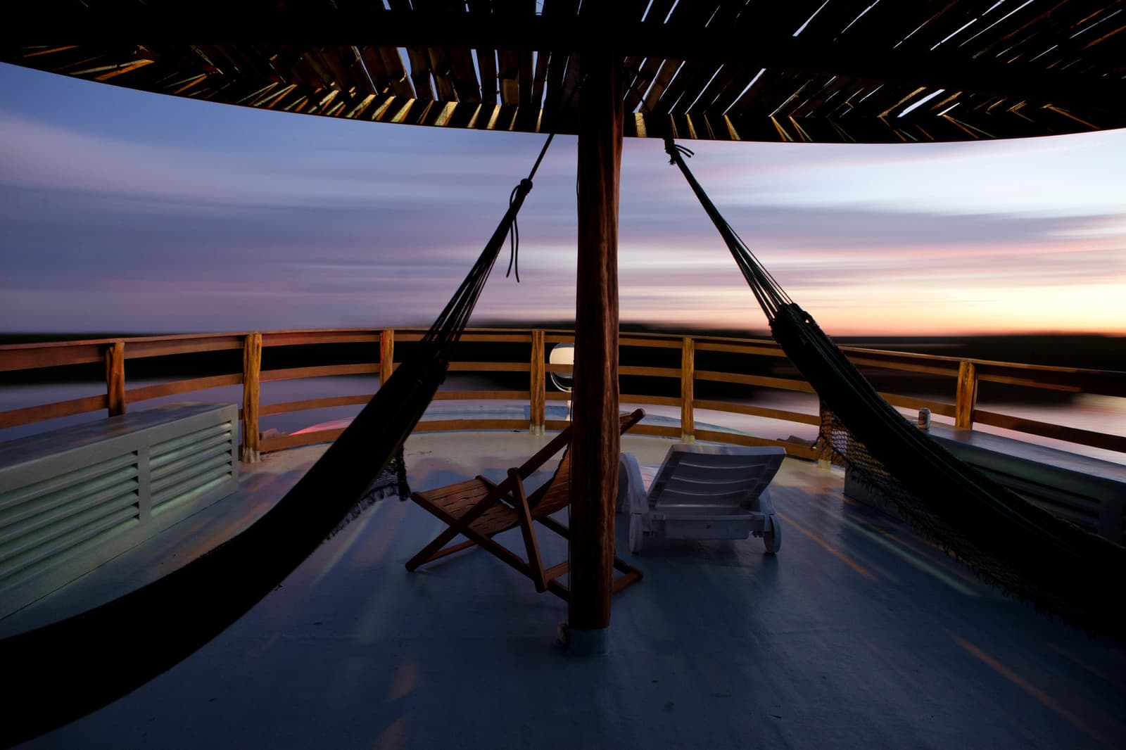 The river decides the pace, and the hammock agrees. Open upper deck of an Amazon river cruise at dusk, with two hammocks hanging from a thatched palapa post, wooden lounge chairs, and a sweeping view of the horizon glowing orange and purple over a flat river landscape