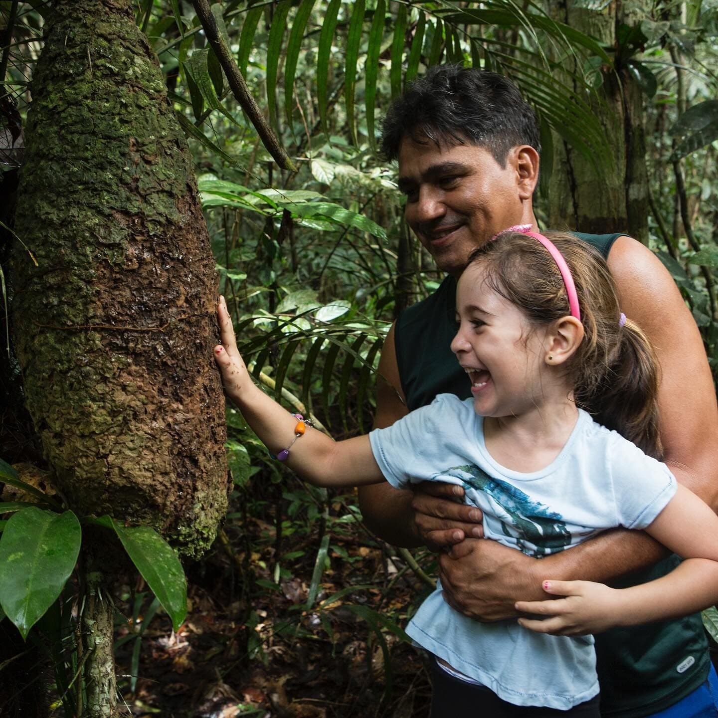 The forest makes a lot more sense when someone who grew up in it shows you around. A laughing girl touches the textured bark of a large rainforest tree during an Amazon jungle cruise Brazil excursion, held gently by a smiling adult guide in lush green forest
