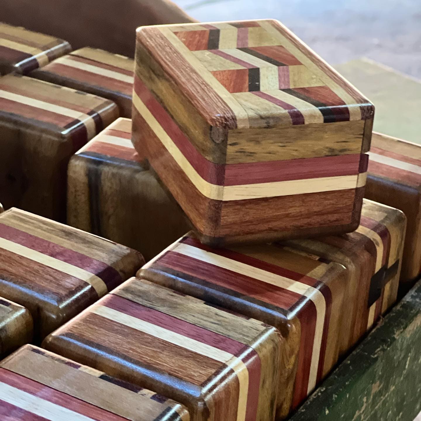Each box holds a different grain, a different story, and no two are quite the same. Collection of small handcrafted wooden boxes with inlaid stripe patterns in warm reds, creams, and dark browns, made from multiple wood species, displayed in a tray during an Amazon river cruise in Brazil stop at a local artisan workshop