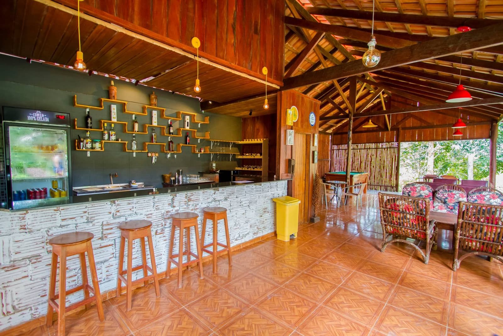 Cold drink, wooden stool, jungle just outside. Not a bad end to a river day. The open-air bar at the Amazon Turtle Lodge features a whitewashed stone counter, wooden barstools, staggered bottle shelves on a dark wall, rattan lounge chairs, and a high timber-beamed ceiling opening onto lush green trees outside.