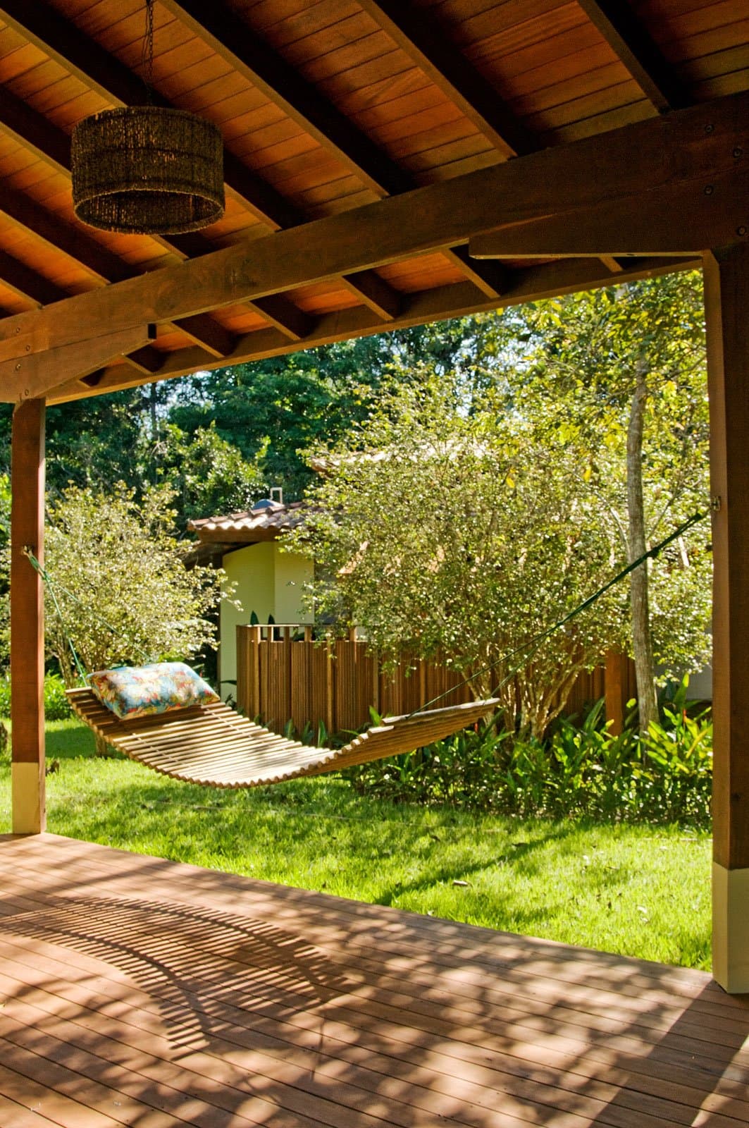 Afternoon light, a slatted hammock, and nowhere to be until the next trail. Photo: Luis Gomes A wooden slatted hammock with a floral pillow rests under a timber-beamed pergola on a hardwood deck, framing a lush green garden and a small lodge building beyond, at a birdwatching in the amazon rainforest retreat
