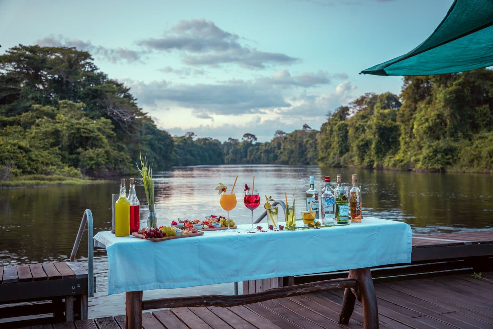Sundowners on the dock, the river quiet, the forest leaning in. Photo: Samuel Melim A white-clothed table on a wooden riverside deck set with tropical cocktails, fruit and cheese boards, and spirits bottles, with dense Amazon rainforest lining a calm river channel at dusk