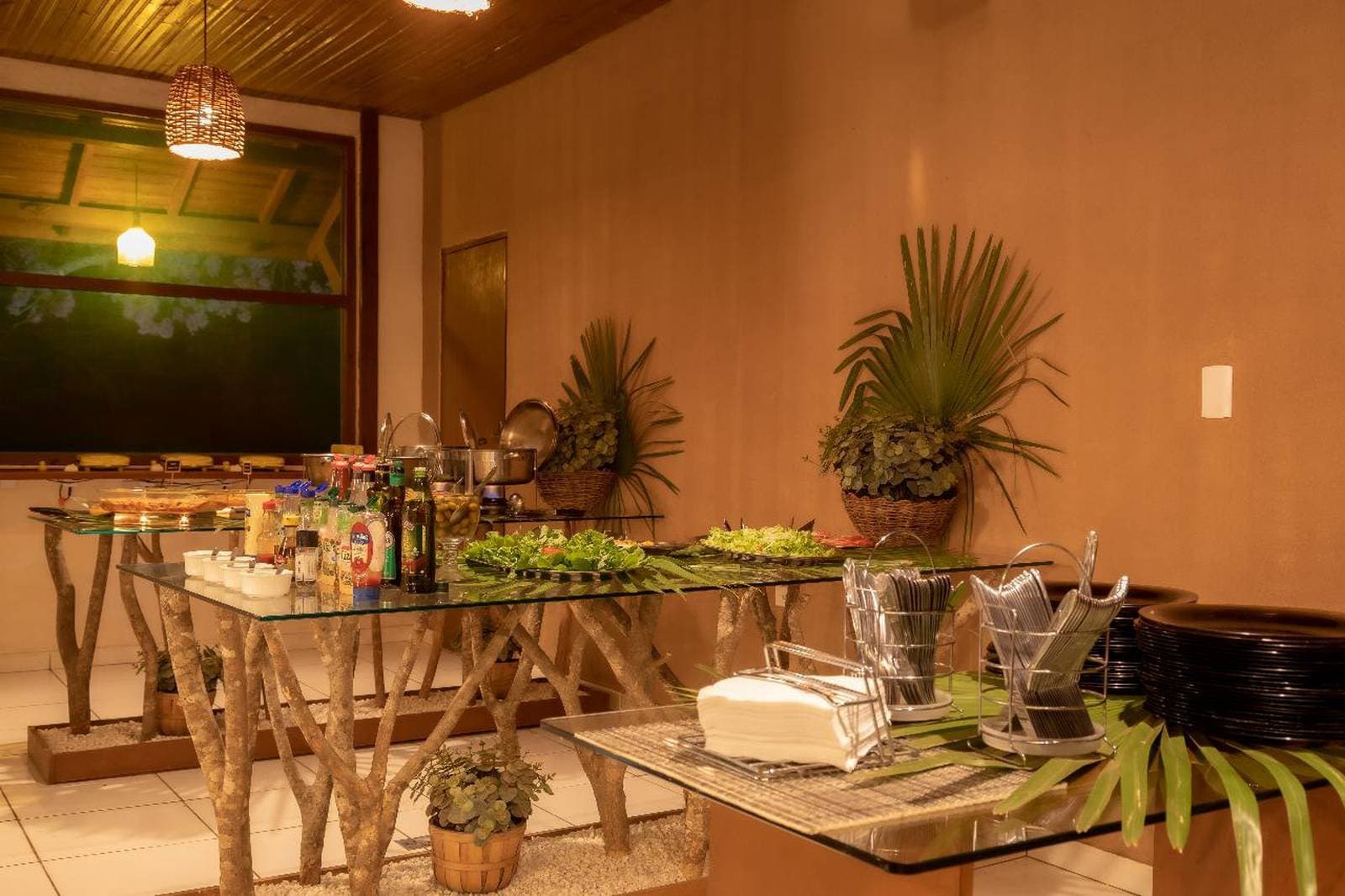 Good food after a long day in the field hits differently. Indoor buffet spread at a Pantanal budget lodge, with glass-topped tables supported by rustic branch legs, loaded with salads, condiments, hot dishes in steel pots, and tropical plant decorations under warm pendant lighting