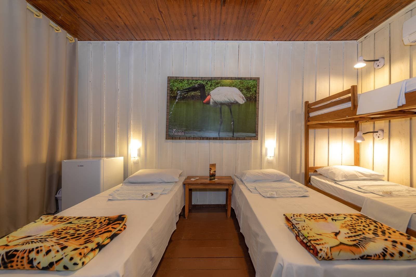 Jaguar blankets, a jabiru on the wall, and a good night's sleep sorted. Cozy multi-bed room at a budget Pantanal tour lodge, featuring two single beds with jaguar-print blankets, a wooden bunk bed, warm wall lights, timber ceiling, and a framed jabiru stork photo on the white panelled wall