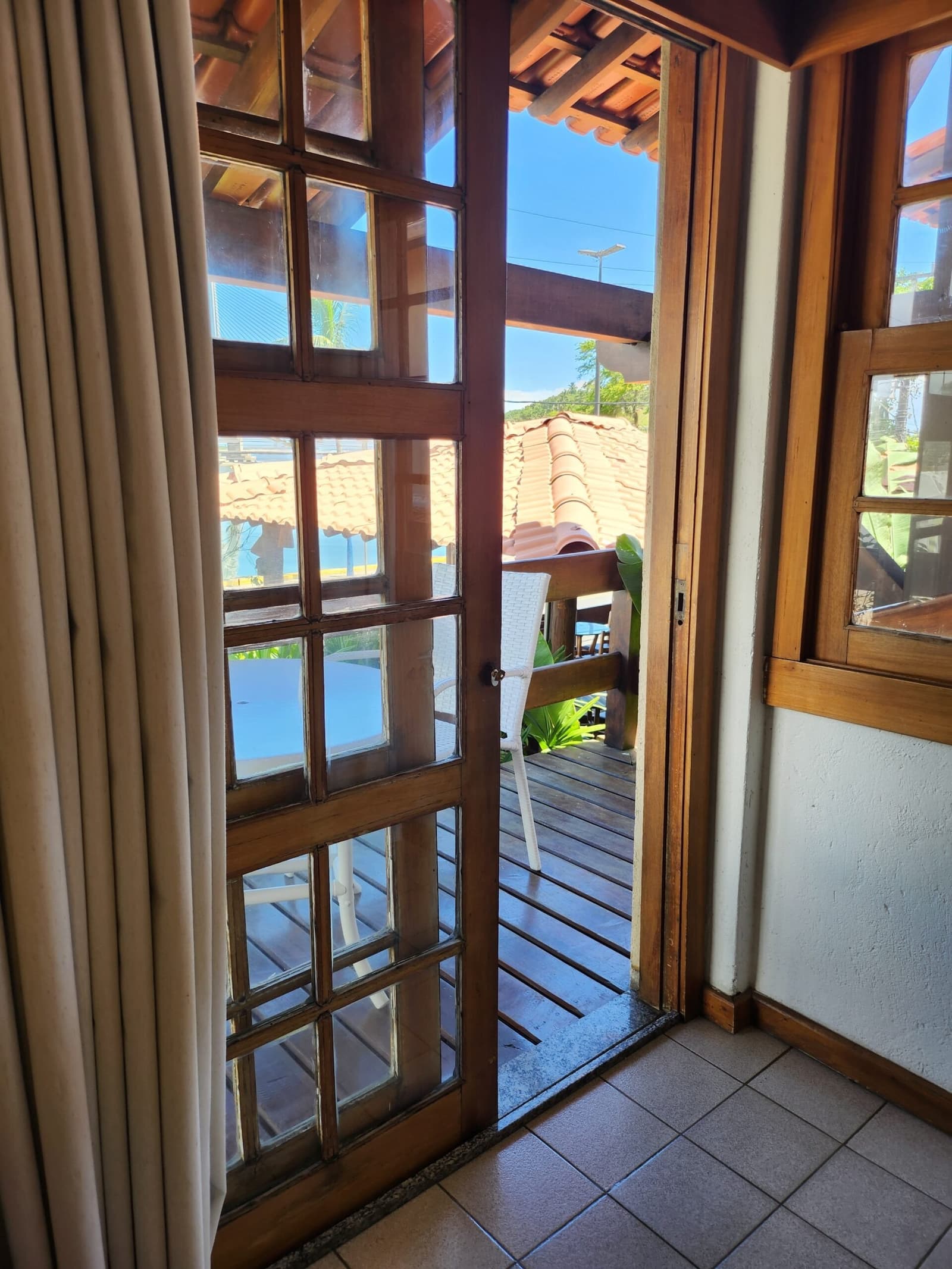 The door is already open. The deck is waiting. Take a minute. View through an open wooden-framed glass door onto a sunlit wooden deck with a white chair, terracotta roof tiles and tropical greenery, at a guesthouse on a cycling bahia tour