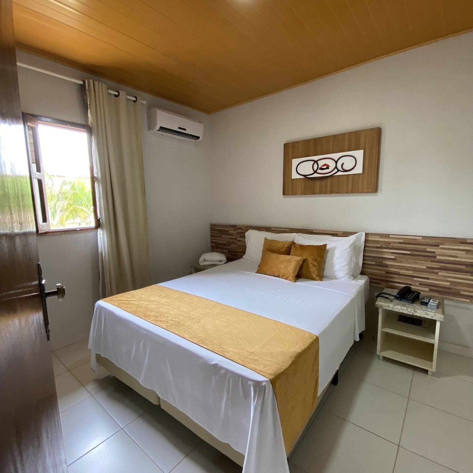 Crisp sheets and a cool room, earned after a day on the saddle. Neatly made double bed with gold accent cushions and bed runner in a clean, air-conditioned hotel room with a wooden ceiling and a window overlooking tropical greenery, part of a cycling Bahia itinerary based in Ilhéus
