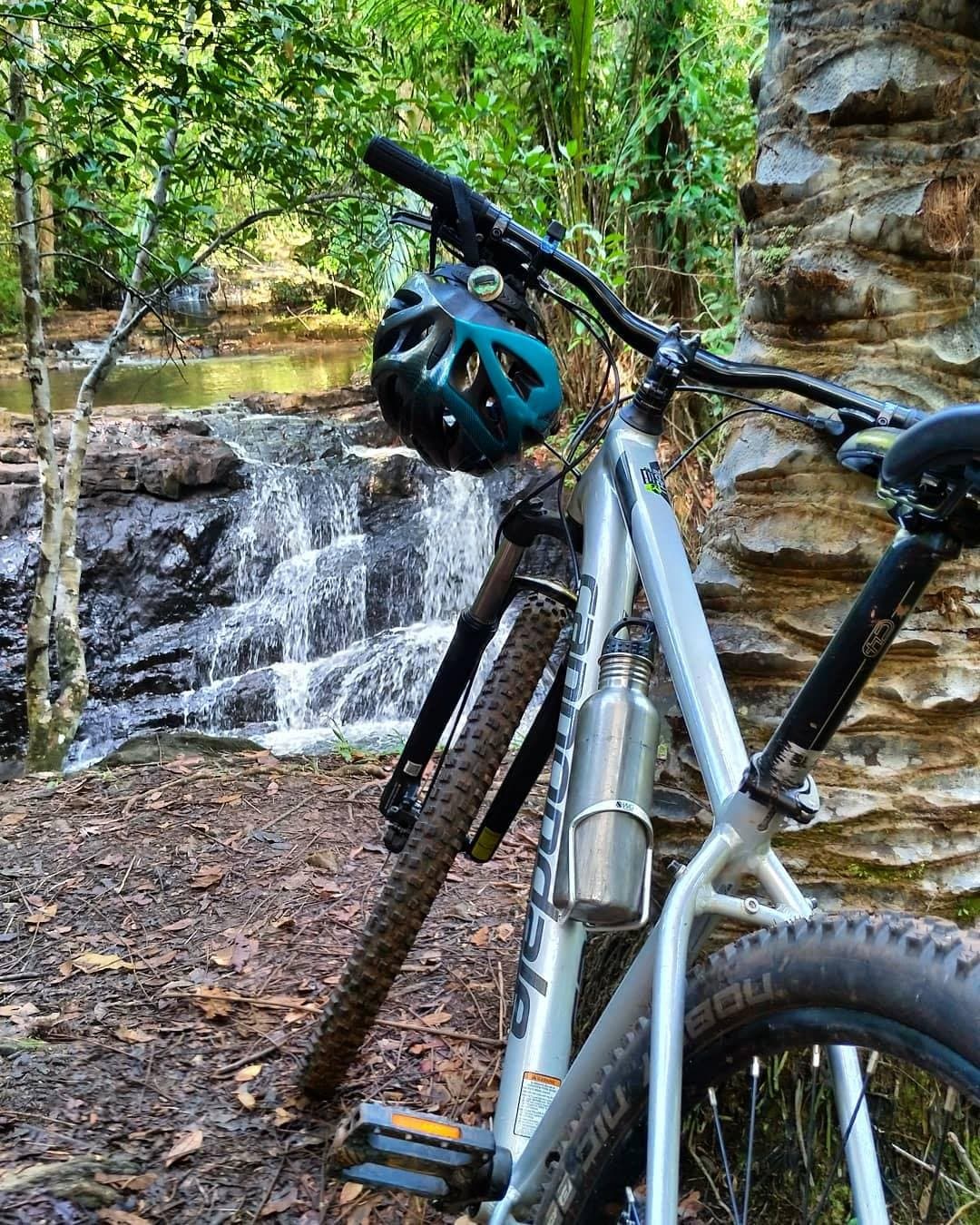 Some stops write themselves. Bike down, helmet off, waterfall on. A mountain bike with a teal helmet resting against a sandstone rock beside a small cascading waterfall on a cycling bahia trail through dense Atlantic Forest