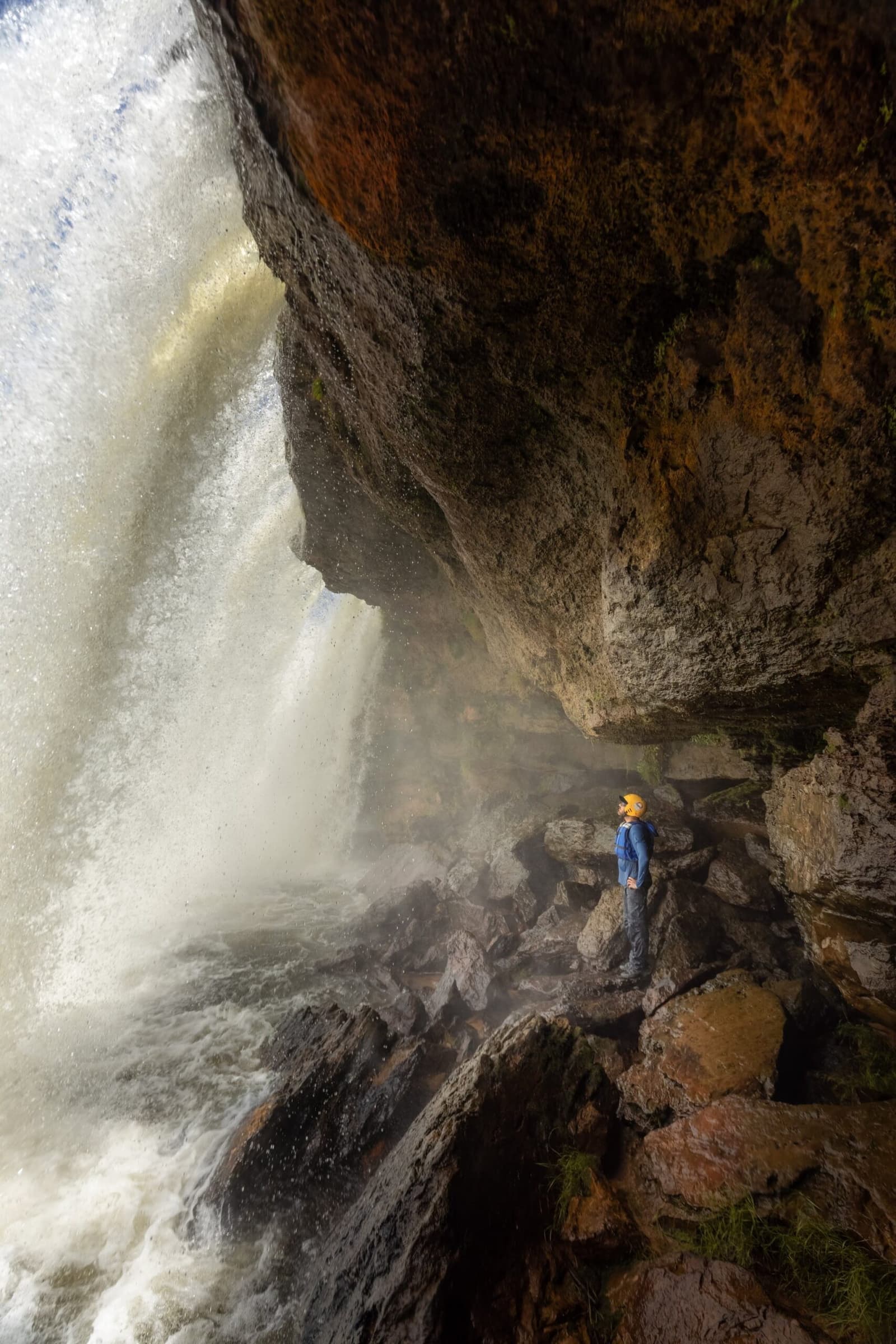 That roar you feel in your chest before you even see the falls. Photo: Alexandre Socci A helmeted hiker in a blue jacket stands on a rocky ledge beneath a thundering waterfall during a Jalapão adventure, surrounded by mist and rust-coloured canyon walls