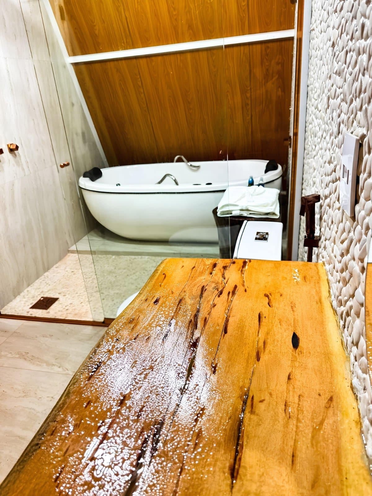Dusty trails, cold springs, then this. The body knows what it needs. A white oval soaking tub set against warm wood-panelled walls in a lodge bathroom, with a wet live-edge wood bench in the foreground and a textured pebble-stone accent wall, part of a Jalapão adventure itinerary