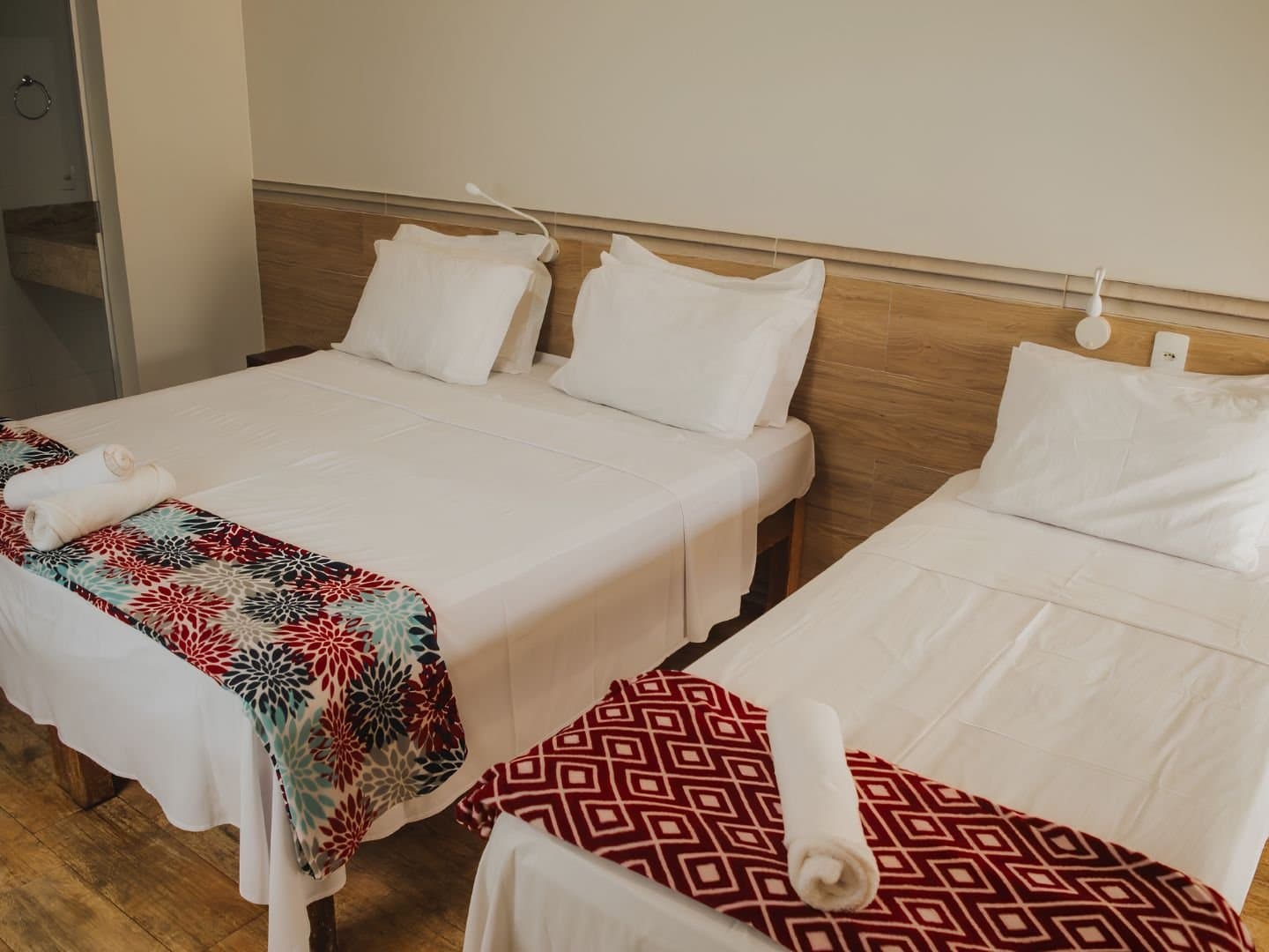 Three beds, fresh towels, and no complaints after a day on the trail. Triple room in a Jalapão adventure guesthouse with two double beds and a single bed, white linens, colorful floral and geometric bed runners, rolled towels, and a wood-panel headboard wall