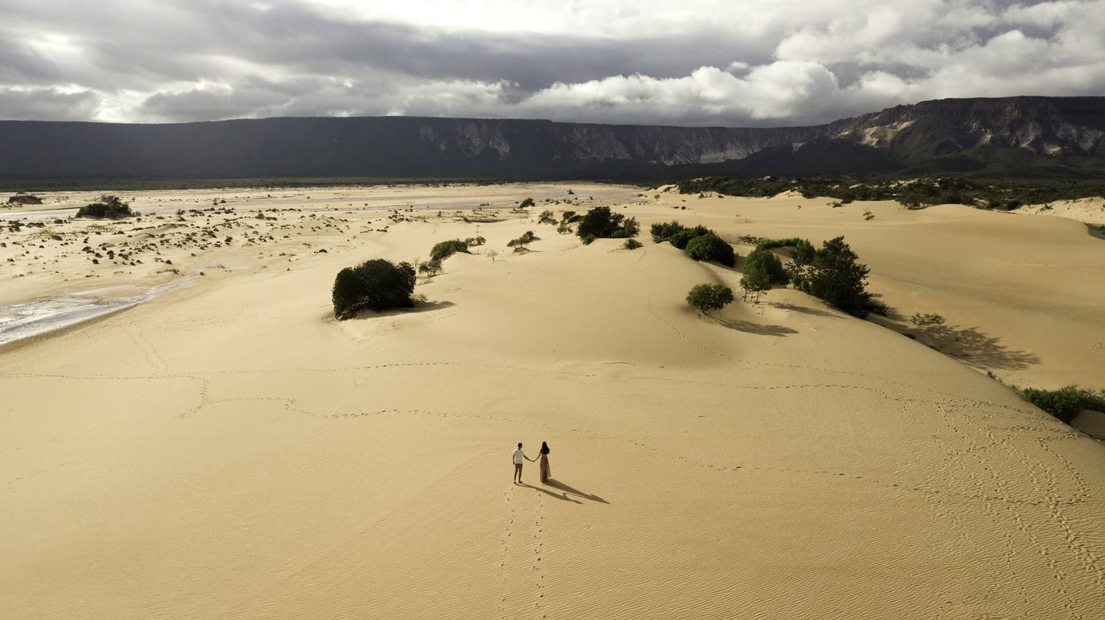 Two people, one enormous dune, and footprints that will be gone by tomorrow. Photo: FG Trade Aerial view of a couple holding hands on the golden sand dunes of Jalapão trip, with green shrubs, a river, and a dramatic sandstone escarpment under a cloudy sky