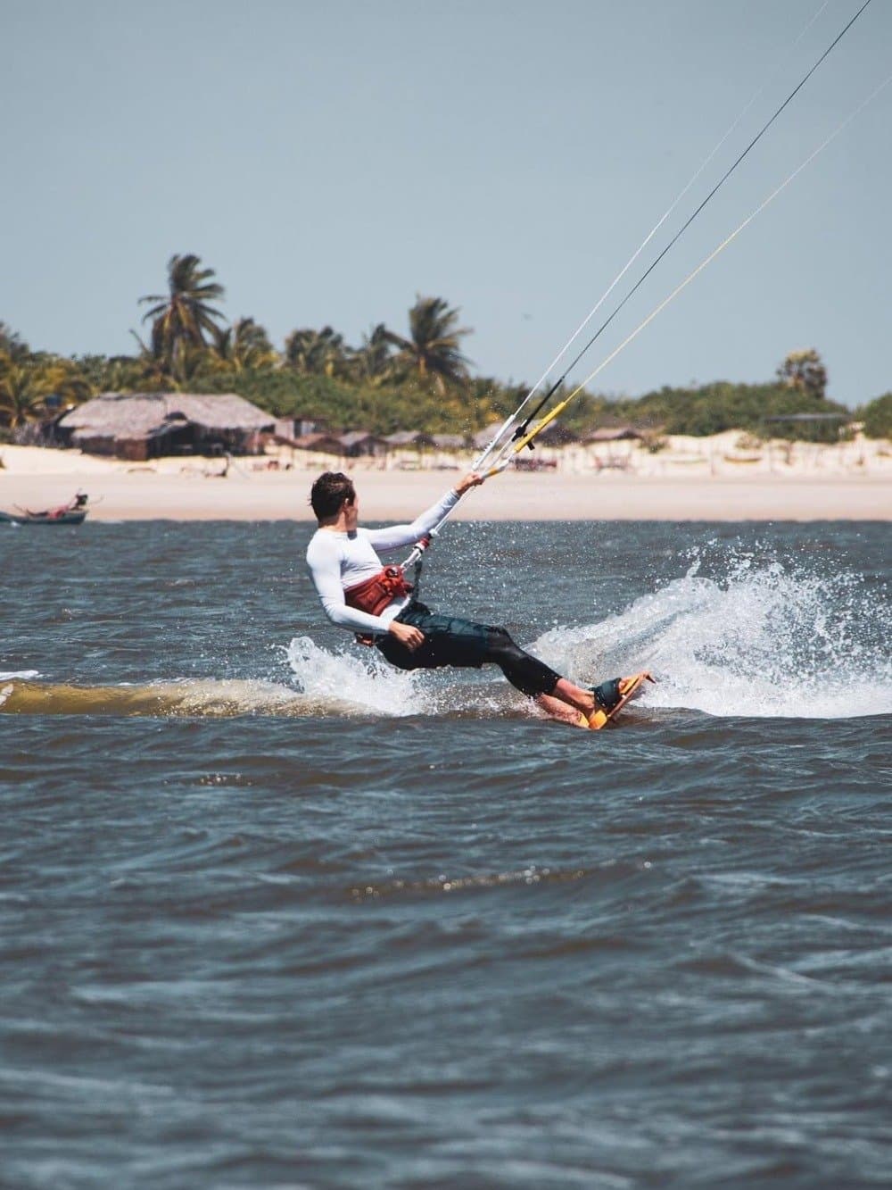 The wind here doesn't ask twice, it just pulls you in. A kitesurfer skims across choppy water near a white-sand beach lined with palm trees and thatched huts, part of the Lençóis Maranhenses full itinerary experience