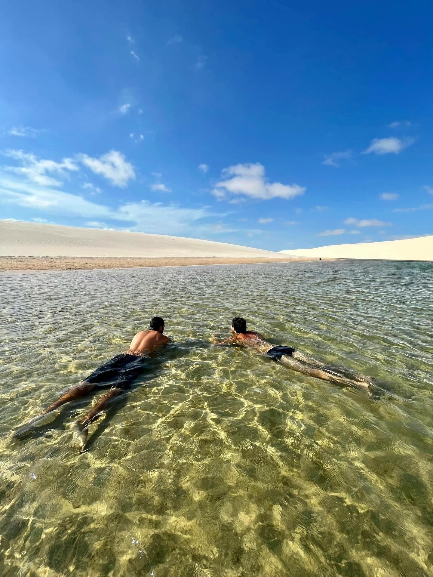 The water is so clear you can see every ripple on the sandy floor beneath you. Two swimmers float face-down in a crystal-clear shallow lagoon on the Lençóis Maranhenses full itinerary, with sweeping white sand dunes stretching to the horizon under a vivid blue sky