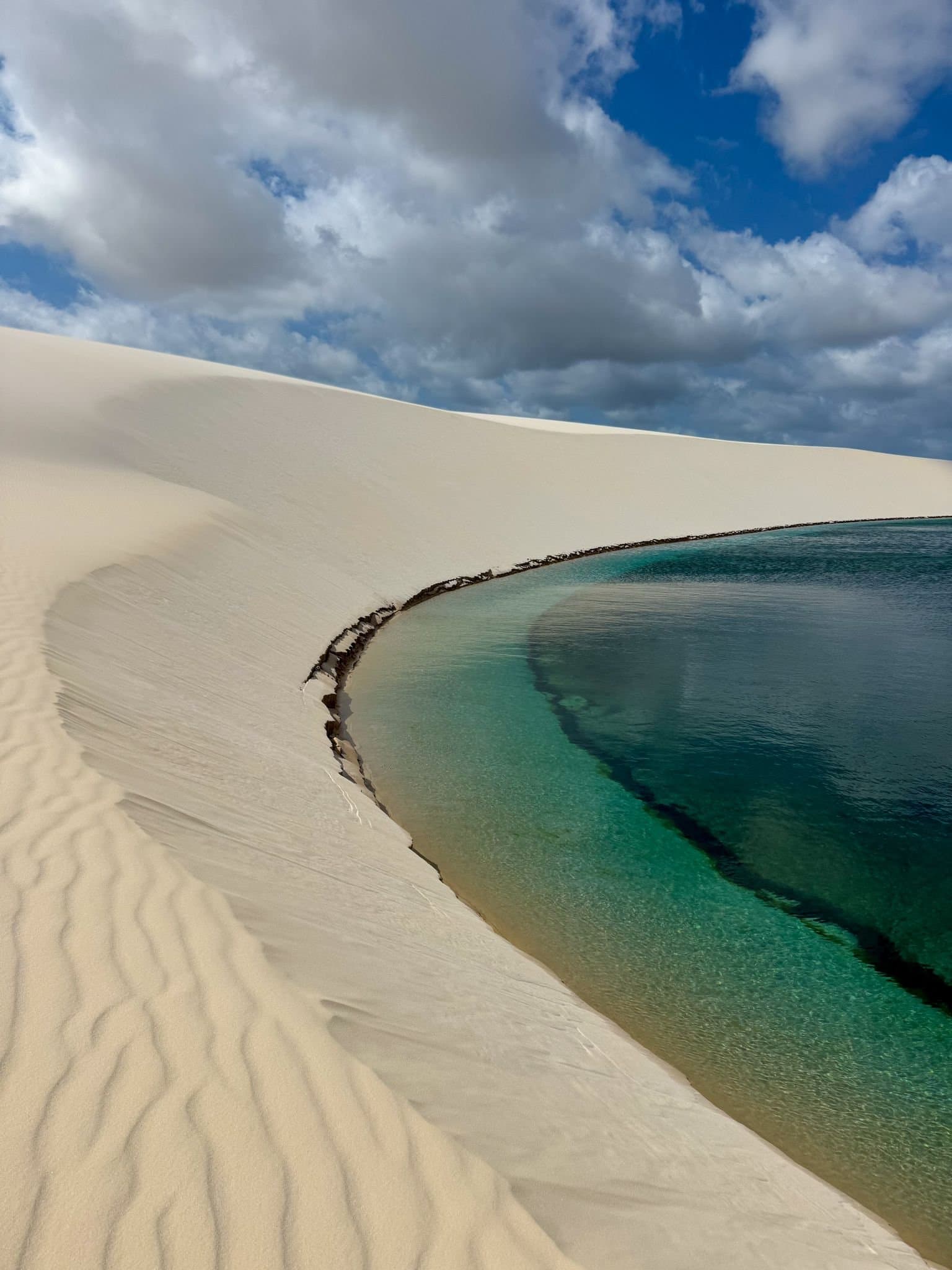 Sand this white and water this clear should not coexist, and yet here we are. A crystal-clear turquoise lagoon nestled against a sweeping white sand dune under a dramatic cloudy sky, part of the Lençóis Maranhenses Full Itinerary experience