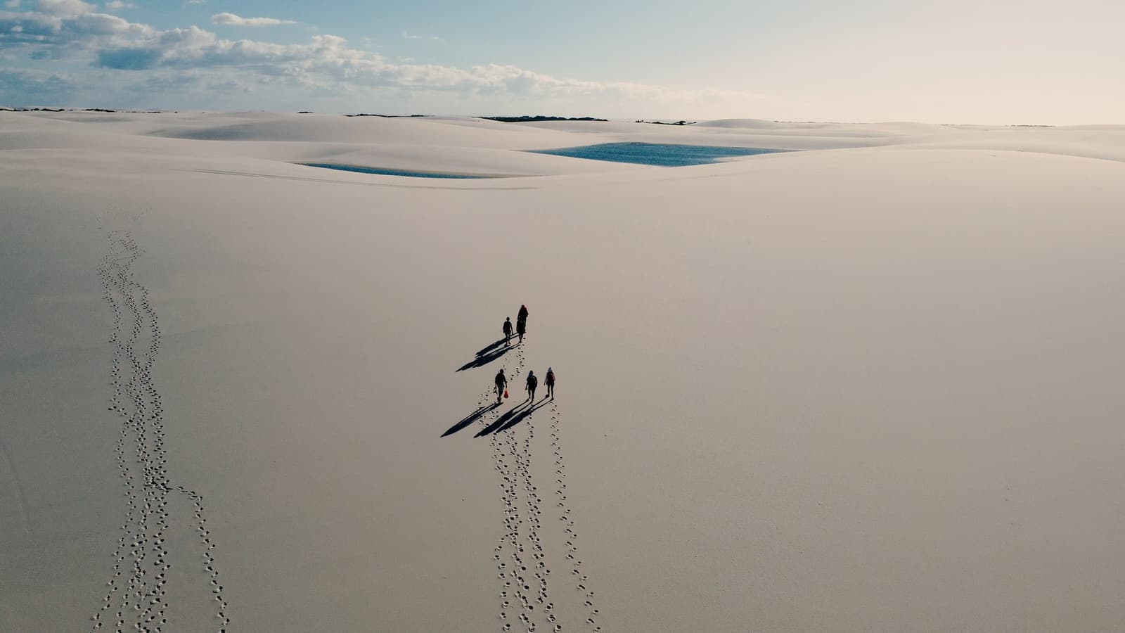 Four people, a whole lot of sand, and the quiet satisfaction of being very small. Aerial view of four hikers walking across vast white sand dunes on a Lençóis Maranhenses hike, their long shadows and footprints trailing behind them, with two blue lagoons visible in the distance