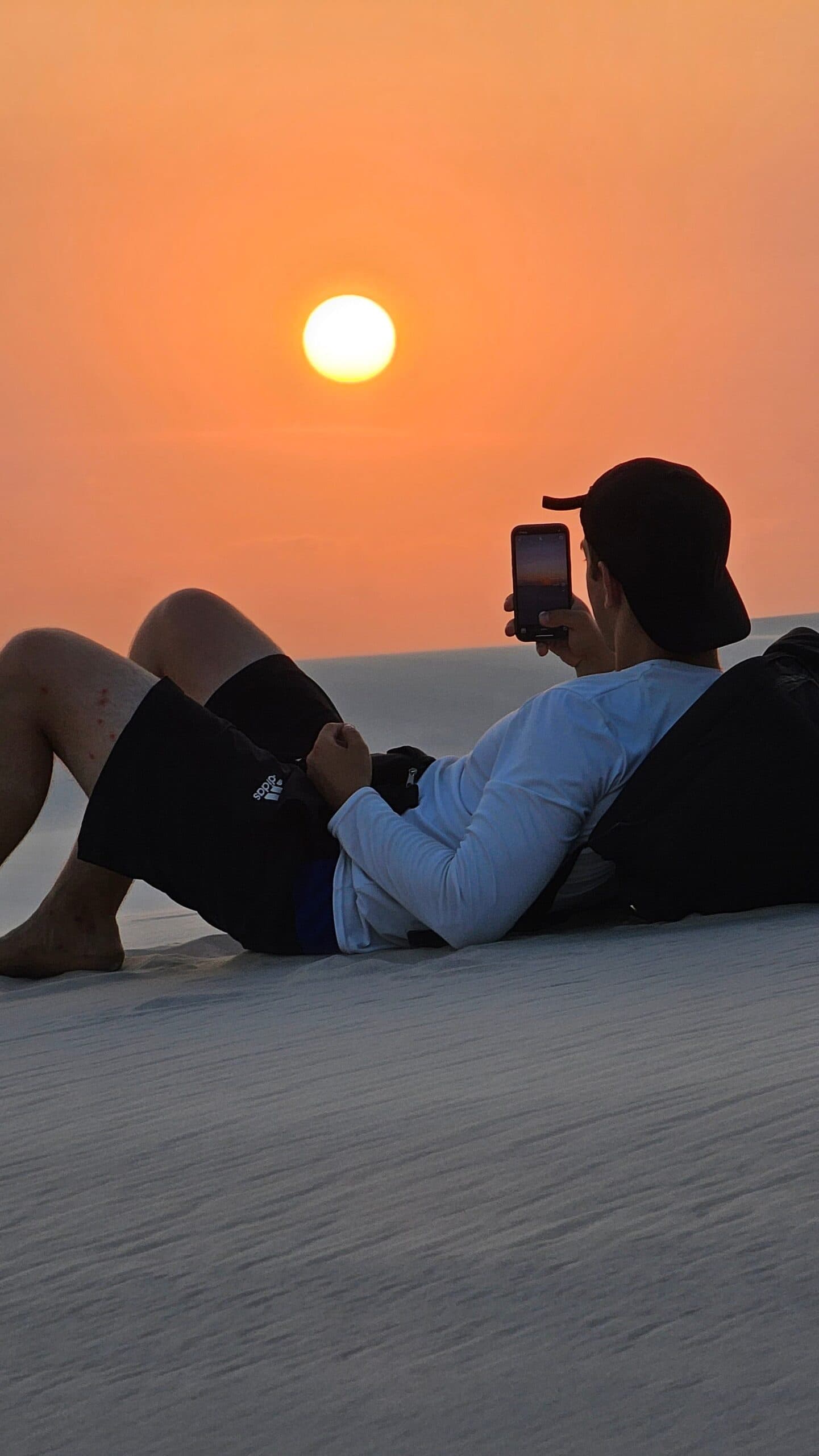 Some sunsets are worth lying down for. A traveller lies back on a white sand dune in Lençóis Maranhenses Santo Amaro, photographing a large orange sun setting over the horizon with their smartphone