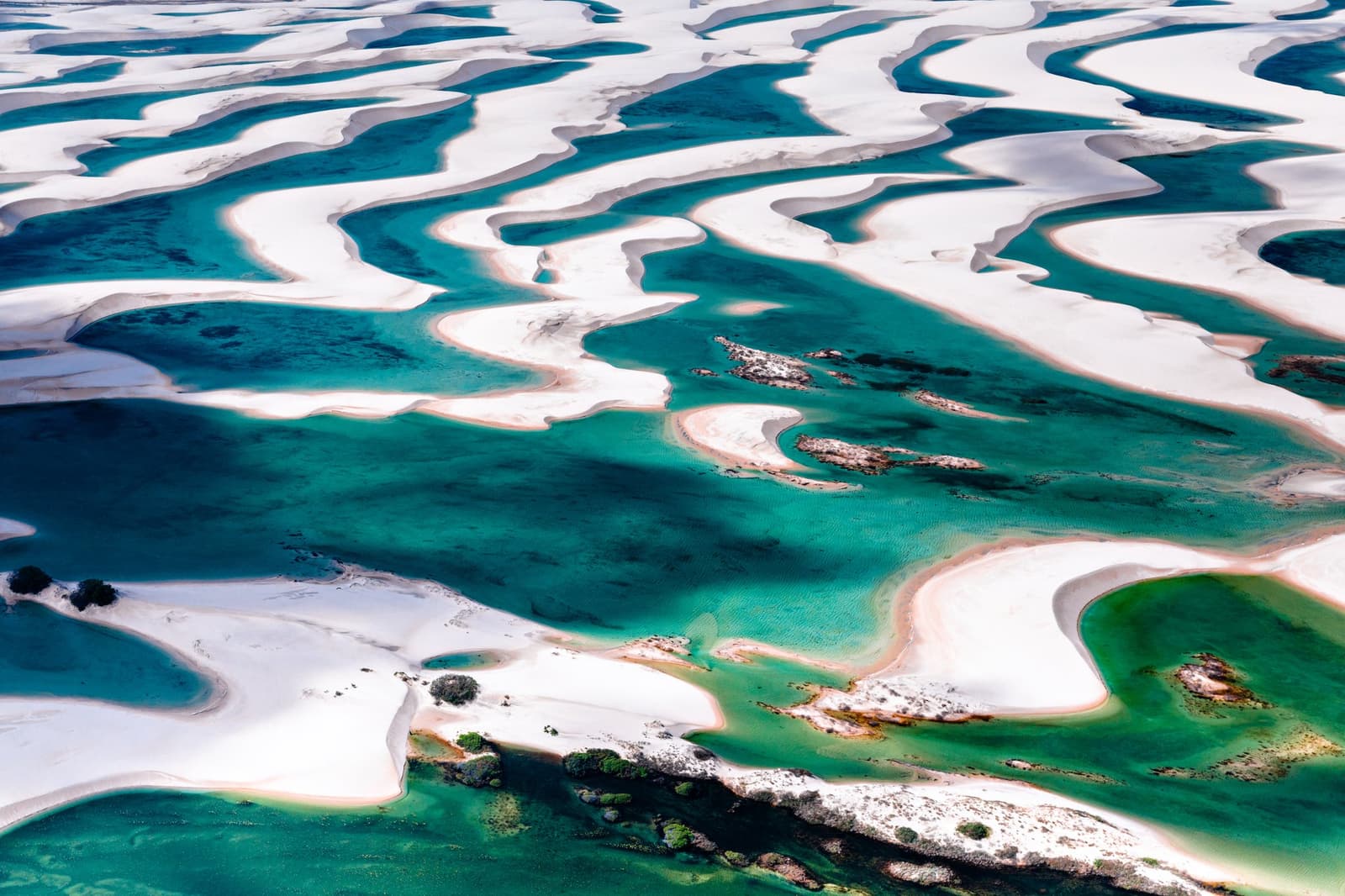 From above, the dunes and lagoons look like something a painter invented on a good day. Aerial view of the lencois maranhenses trek landscape, showing vast white sand dunes interlaced with curving turquoise and emerald freshwater lagoons stretching to the horizon
