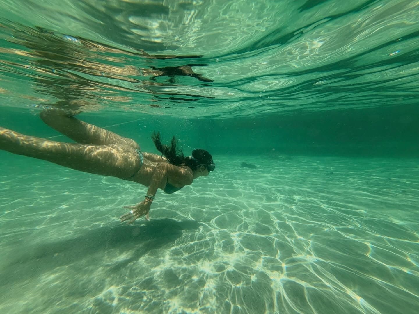 That first dive into a freshwater lagoon resets everything. A woman free-diving underwater in a crystal-clear shallow freshwater lagoon during the Lençóis Maranhenses trek, sunlight rippling across the sandy floor