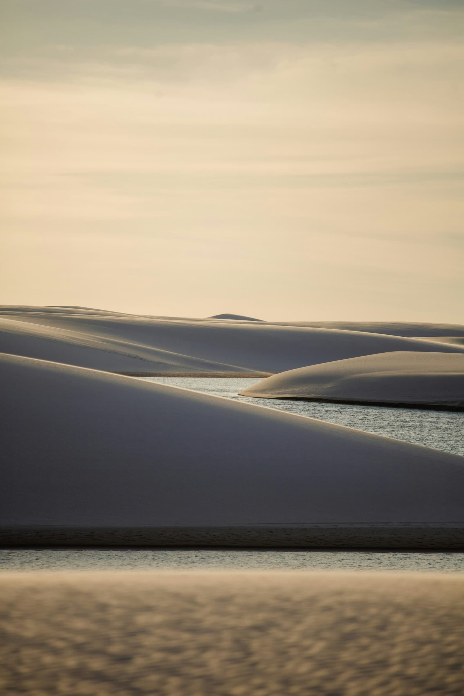 Sand that behaves like sculpture, water that appears from nowhere. Sweeping white sand dunes of the lencois maranhenses trek casting deep blue shadows over a narrow freshwater lagoon at golden hour, with a pale hazy sky above