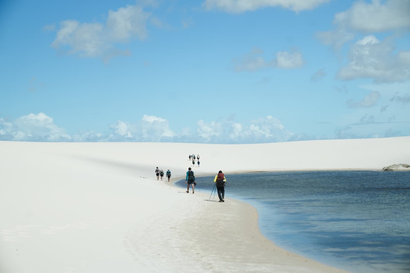 One foot in the dune, one foot in the water, that's the whole trail right there. A group of trekkers with backpacks walking single file along a narrow strip of white sand between a large blue lagoon and sweeping dunes during a Lençóis Maranhenses trekking expedition