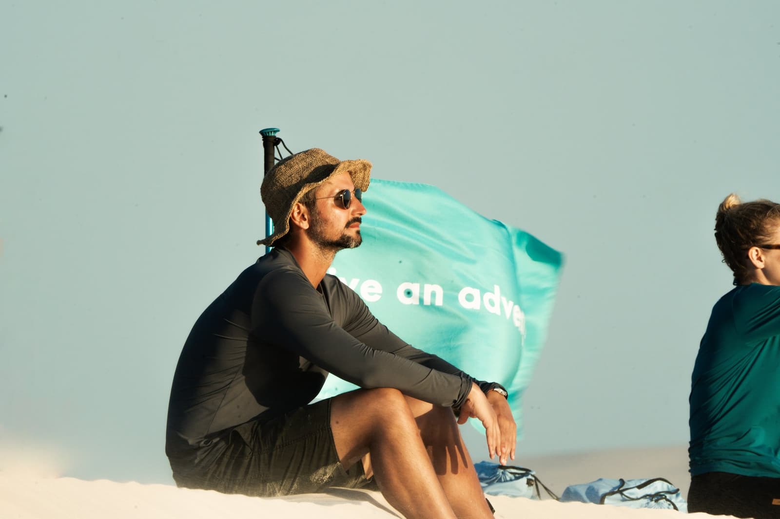 Even guides need a moment to just sit and look at nothing in particular. A man in a straw hat and sunglasses sits on white sand dunes during a Lençóis Maranhenses trekking break, with a turquoise flag flying behind him in the wind
