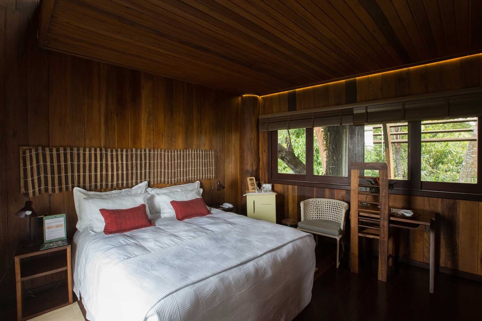 Warm timber walls, forest just outside the glass, sleep is easy here. A Mirante do Gavião amazon lodge guest room with dark timber walls, crisp white bedding, red accent cushions, and windows overlooking lush green rainforest