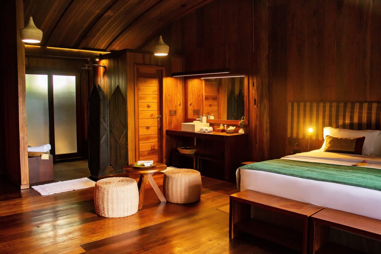 After a day on the river, this is exactly where you want to be. Warmly lit wooden cabin room at Mirante do Gavião amazon lodge, featuring a double bed with green throw, wicker stools, open-plan vanity with mirror, and polished hardwood floors