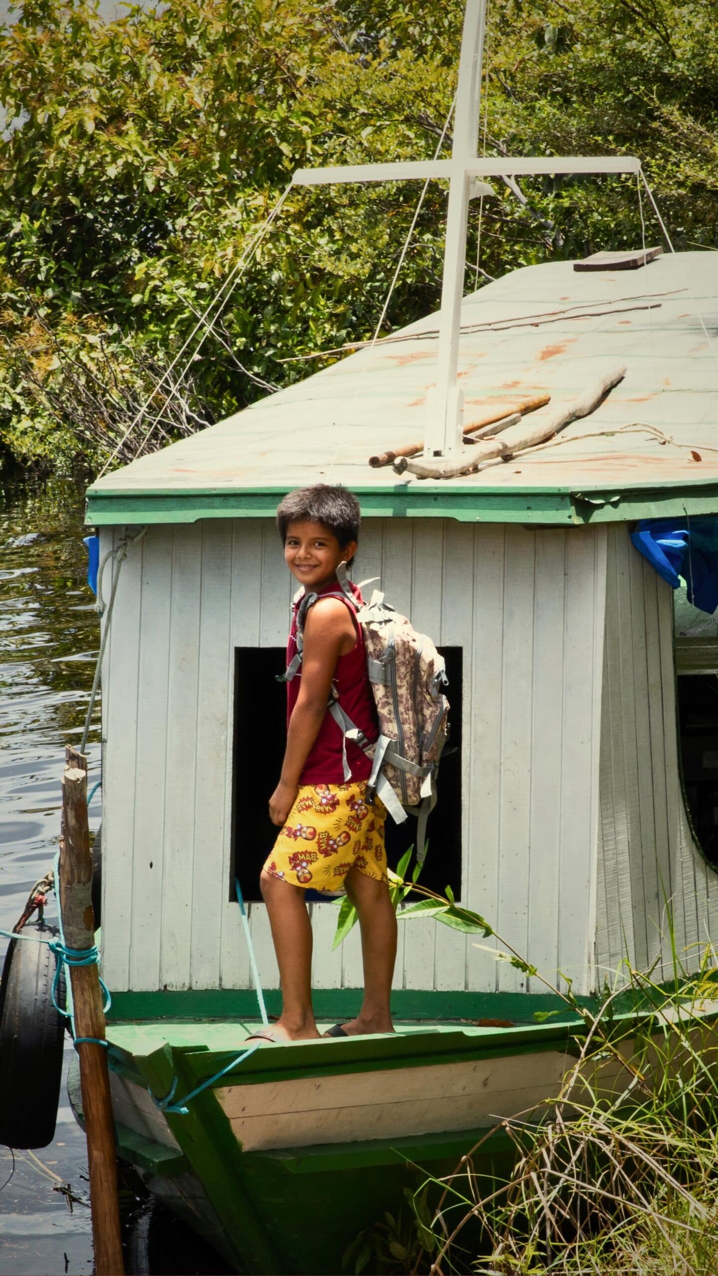 School run, Amazon-style: a backpack, a boat, and no complaints. A smiling boy wearing a backpack stands barefoot on a small wooden houseboat moored along a jungle riverbank, part of a riverside community visit during a Mirante do Gavião tour