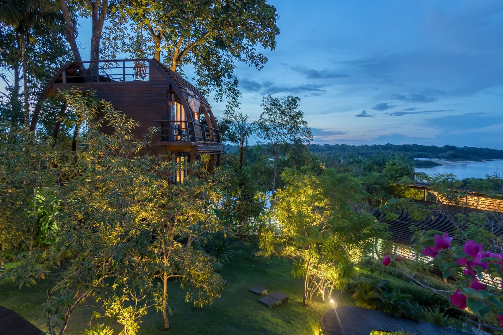 Your room for the night, hovering above the treetops with the river right there. An arched timber treehouse suite at Mirante do Gavião glows with warm interior light at dusk, surrounded by lush tropical garden and overlooking a wide river bend under a blue twilight sky