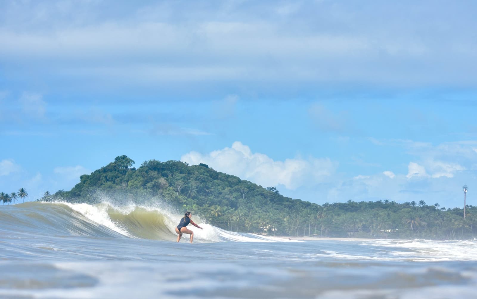 Reading a wave well is half the lesson. The other half feels like this. Photo: Picasa A woman crouches low on her surfboard at the surf school in Itacaré, carving along a peeling wave with a lush forested headland and palm-lined beach in the background
