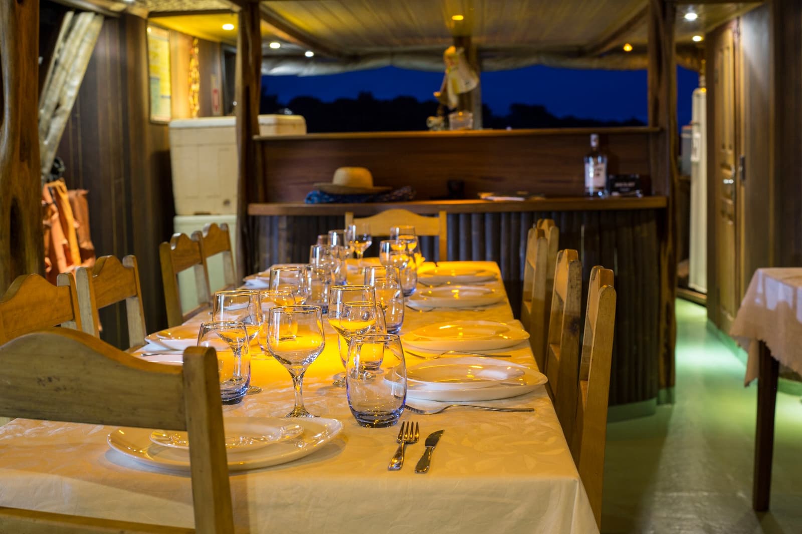 Dinner is ready, the river is waiting, and the night shift belongs to the frogs. A long dining table set for an Amazon cruise trip in Brazil, with white plates, wine glasses, and silver cutlery glowing under warm cabin lights aboard a wooden river boat at dusk