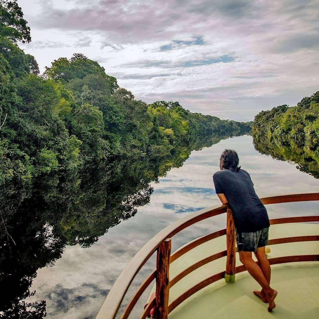 Nothing ahead but forest, water, and the quiet hum of the river. A passenger leans on the wooden bow railing of a riverboat on an amazon river cruise in brazil, gazing down a glassy black-water channel flanked by dense rainforest canopy reflected in the still water