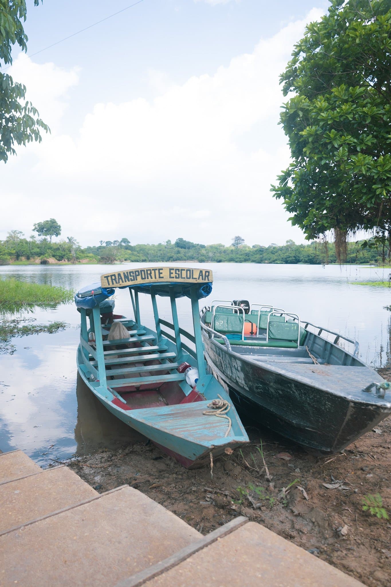 The school run looks a little different out here, and honestly, more fun. Two wooden motorboats moored on a calm river bank near the Amazon Turtle Lodge, one painted turquoise with a 'Transporte Escolar' sign, lush rainforest reflected in the still water behind
