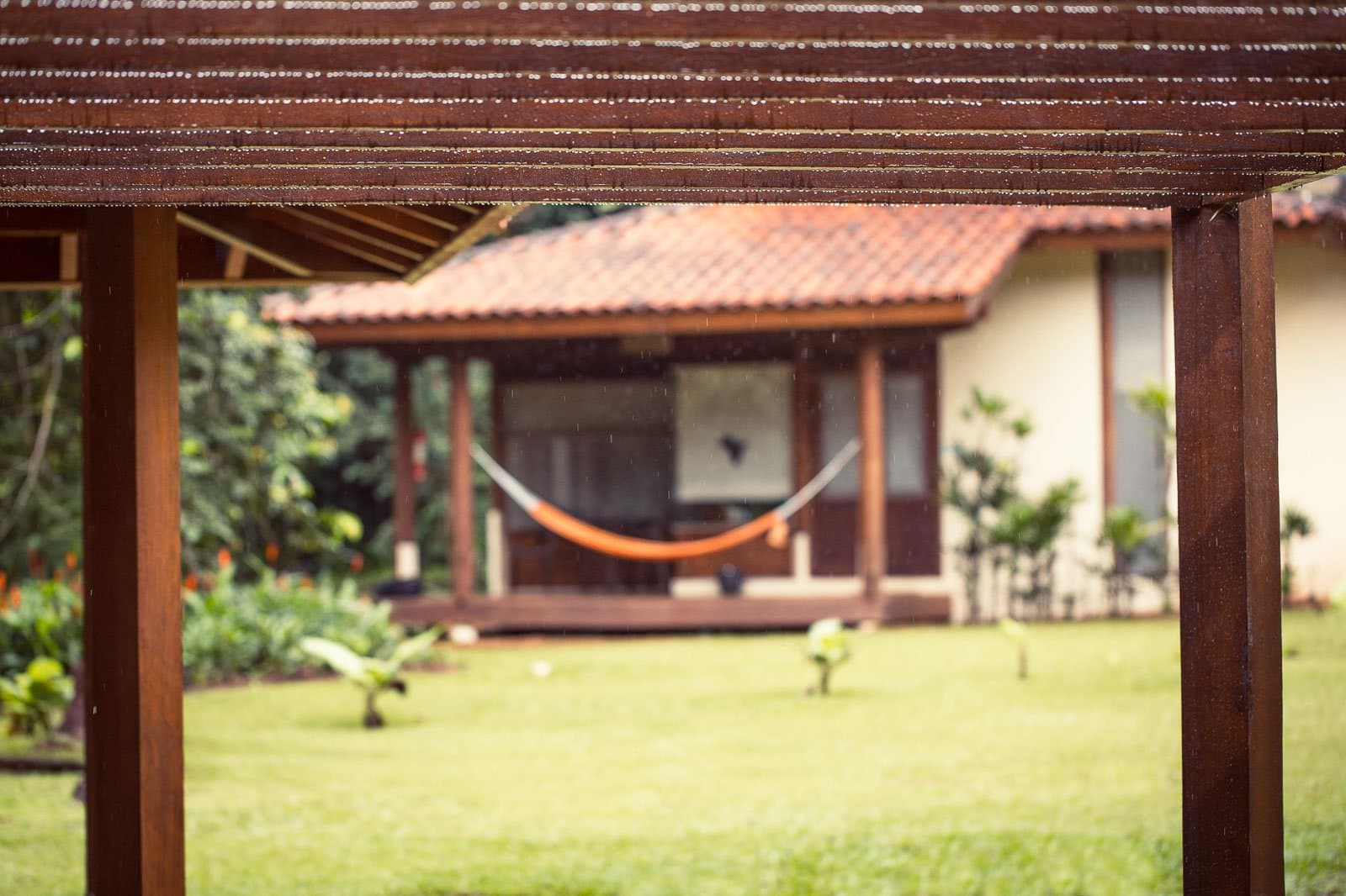 Rain on the roof, hammock waiting. Some afternoons are theirs. Photo: | SamuelMelim Photography A wooden pergola in the foreground frames a view of a jungle lodge bungalow with a terracotta-tiled roof, an orange hammock on the veranda, and a green lawn during a rain shower — the kind of base guests return to between birdwatching in the Amazon rainforest