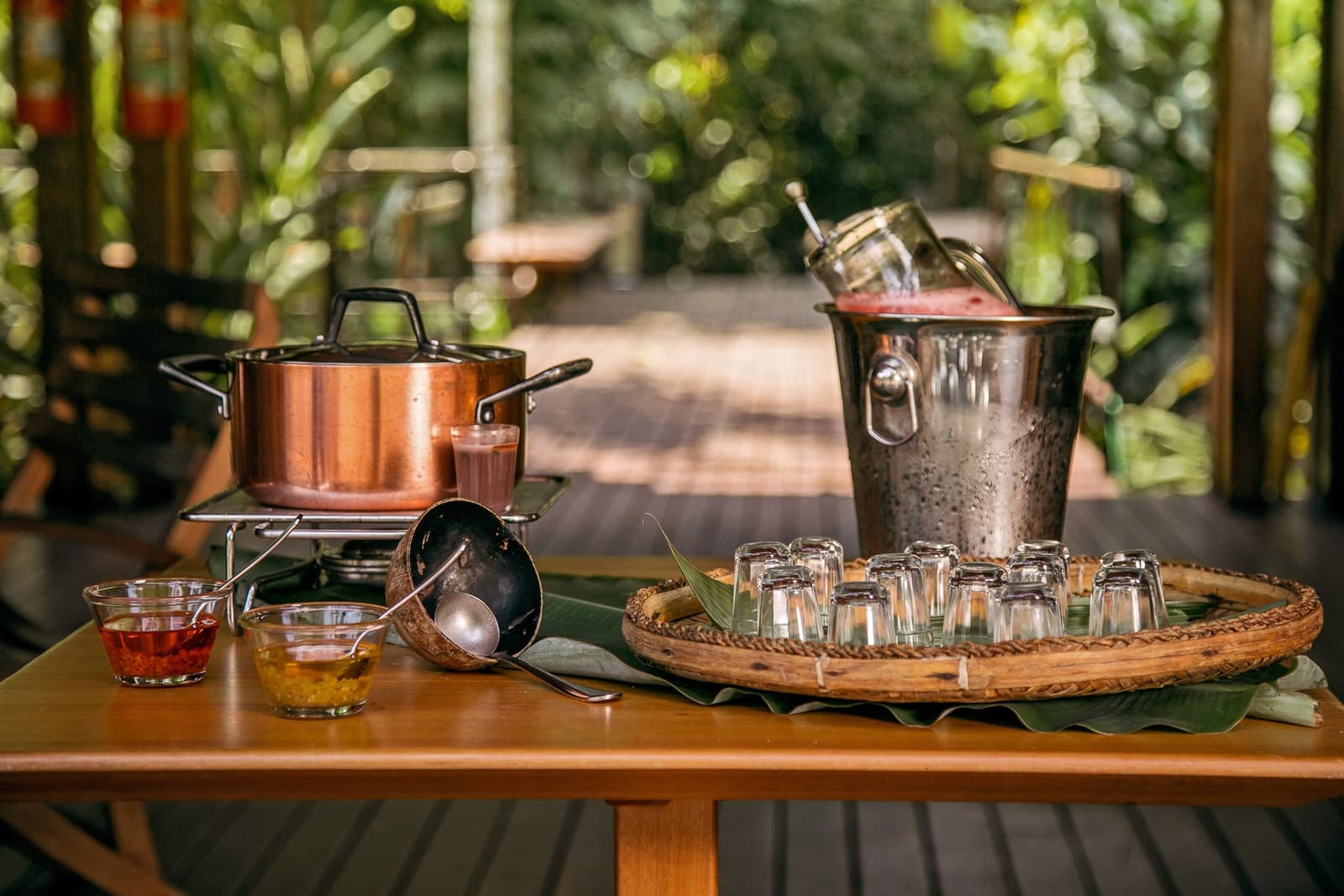 Drinks chilled, pot on the flame. The Amazon has its own idea of a welcome. Photo: | SamuelMelim Photography A copper pot warming on a portable burner beside a hammered silver ice bucket filled with a pink juice and mason jars, small glass shot glasses arranged on a woven tray lined with banana leaves, and two bowls of condiments on a wooden table at an open-air jungle lodge terrace