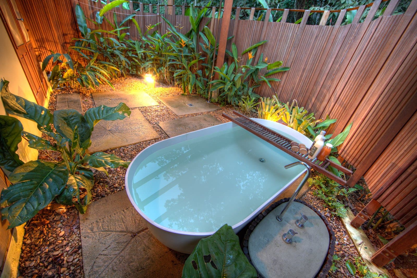 This is what you come back to after four hours on the trail. Photo: Samuel Melim A freestanding white soaking tub filled with water sits in a private open-air garden courtyard, surrounded by tropical foliage, pebble paths, stone pavers, and warm ground lighting enclosed by wooden slatted fencing — part of a birdwatching in the Amazon rainforest lodge experience