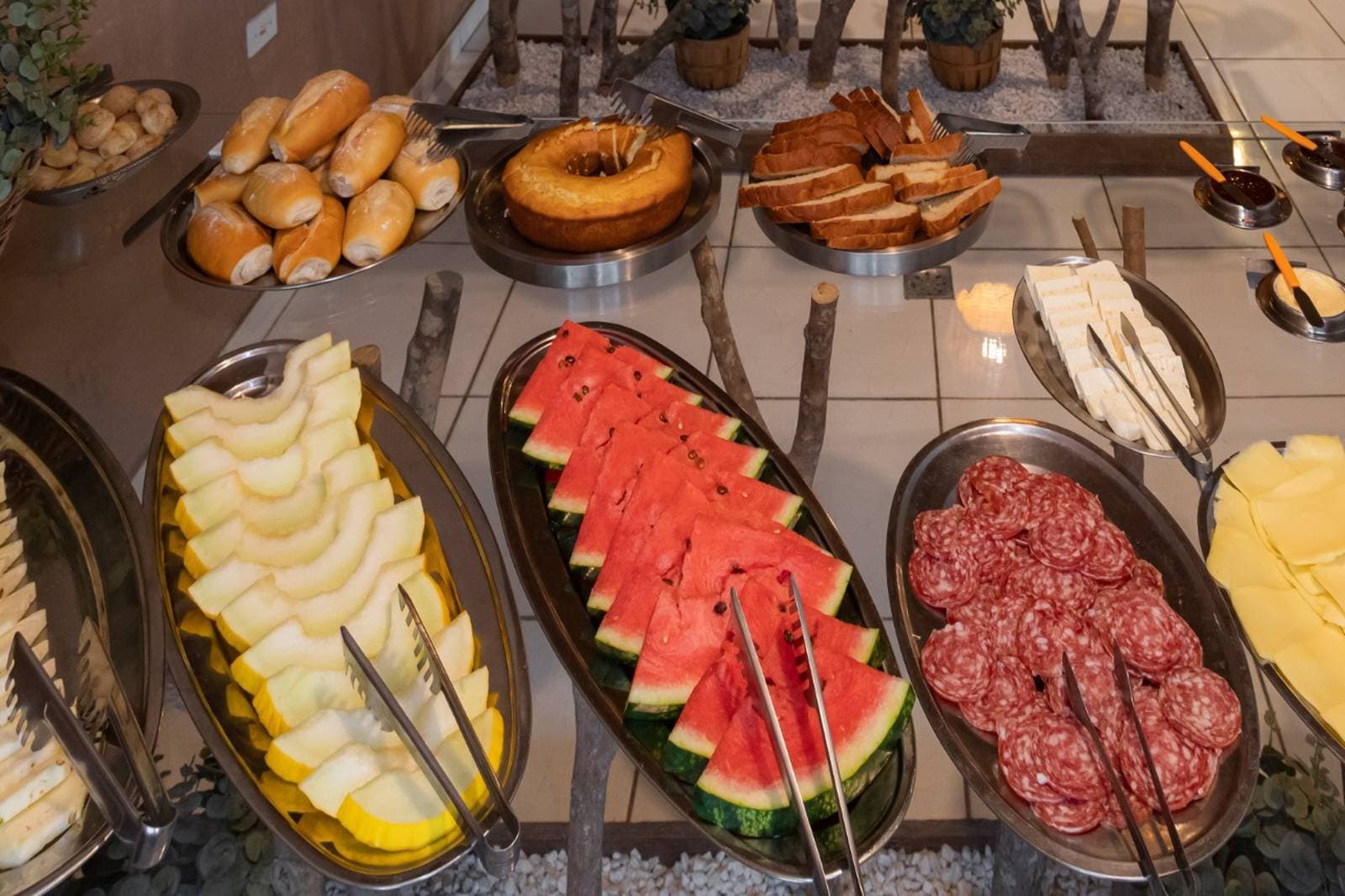 Fuel up well, the wildlife is not going to wait. A full breakfast buffet spread on a budget pantanal tour, featuring sliced watermelon, honeydew melon, salami rounds, sliced cheese, bread rolls, a ring cake, toast, and dipping sauces arranged in stainless steel trays
