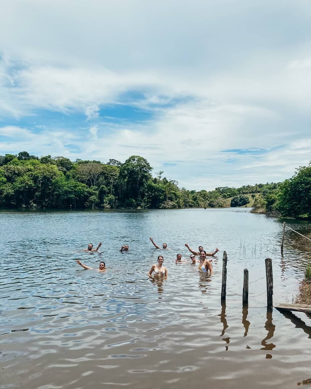 Lunch earned, bikes resting, everyone in the water at once. A group of cyclists swimming and waving in a calm lake surrounded by dense green forest on a bahia cycling trip, with wooden fence posts visible at the water's edge