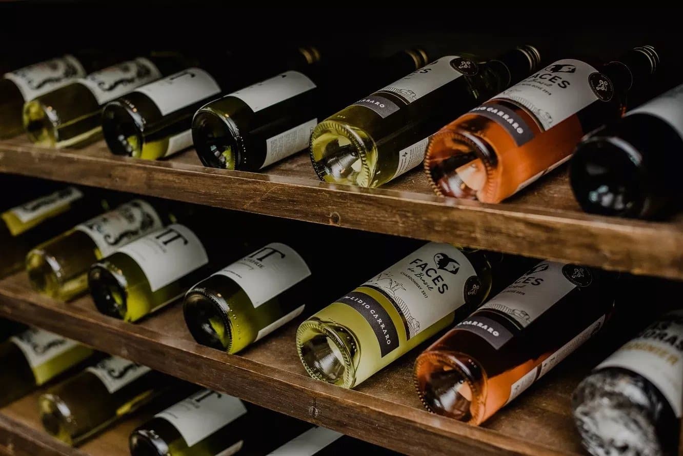 A well-stocked rack is its own kind of welcome at the end of a long day. Bottles of Lidio Carraro Faces do Brasil Chardonnay and rosé wines resting on wooden cellar racks, part of a cycling Bahia tour's evening dining experience