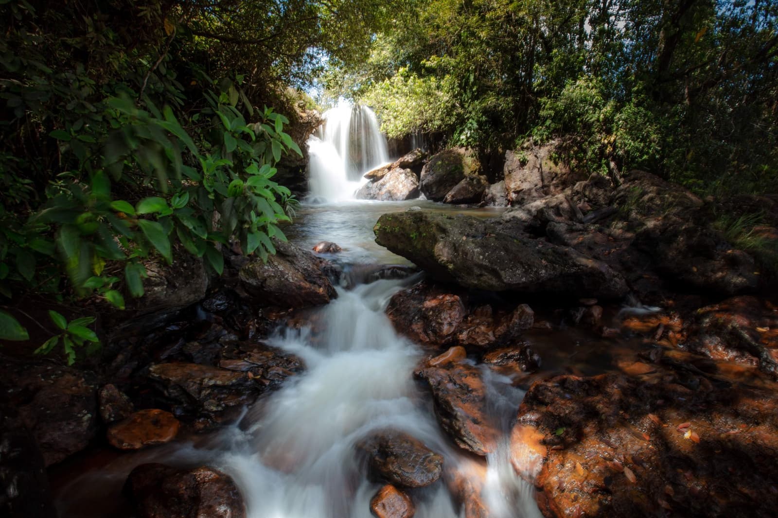 Cool water, warm rocks, and a stream that has absolutely no plans to slow down. Photo: Alberto Andrich A silky waterfall drops through dense tropical forest onto moss-covered rocks, feeding a fast-moving stream over rust-coloured boulders on a Jalapão adventure trail