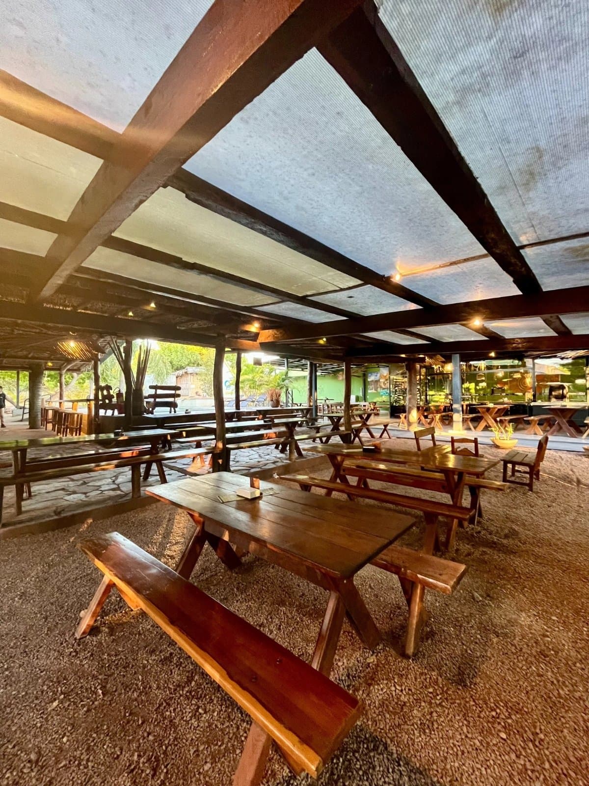 Good food tastes better when the walls are optional. Open-air rustic dining area with rows of polished wooden picnic tables and benches under a shade canopy supported by dark timber beams, with greenery visible at the edges, part of a jalapão adventure guesthouse