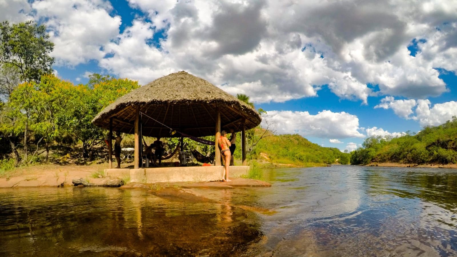 A hammock in the shade, a river at your feet. Not a bad reward for the trail. A thatched open-air shelter with hammocks sits on a low stone platform beside a clear, shallow river in Jalapão, with green cerrado hills and a dramatic cloudy sky behind it; a traveler in a bikini stands at the edge looking out over the water
