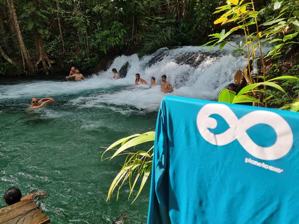 Cold water, dense canopy, and absolutely no reason to get out yet. A group of travelers swimming and wading at the base of a cascading waterfall surrounded by dense tropical forest during a Jalapão trekking tour, with a PlanetaEXO branded towel hanging in the foreground