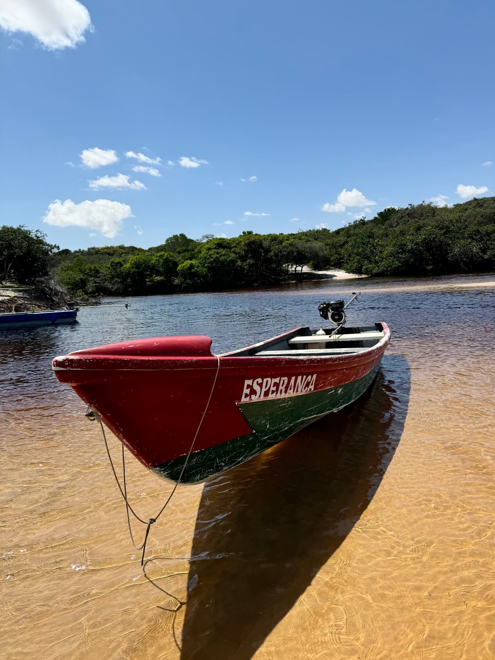 Esperança, hope, tied up and ready, as all good river boats should be. A red and green wooden boat named Esperança moored at a sandy riverbank, with mangrove-fringed water and blue sky behind — part of the Lençóis Maranhenses full itinerary river experience