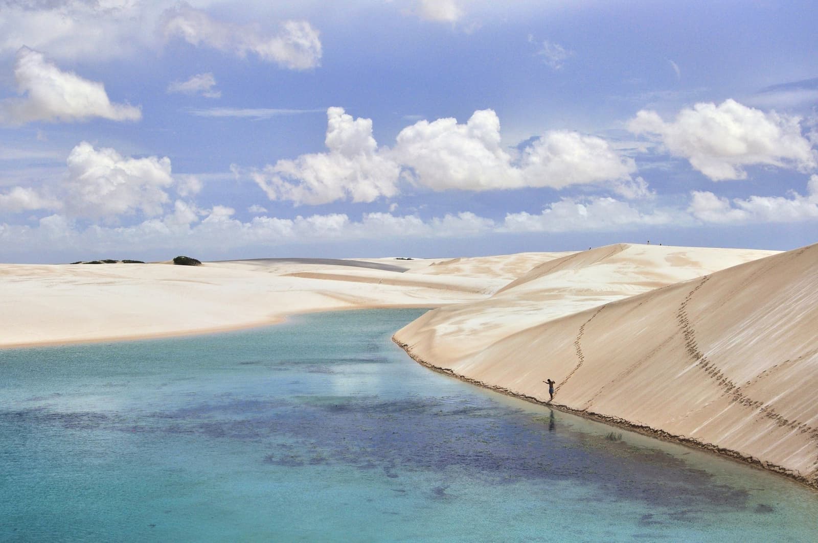 One person, one lagoon, and a whole lot of sand between them and the rest of the world. A lone figure wades along the base of a sweeping white sand dune beside a turquoise lagoon in Lençóis Maranhenses Full Itinerary country, under a wide blue sky with cumulus clouds
